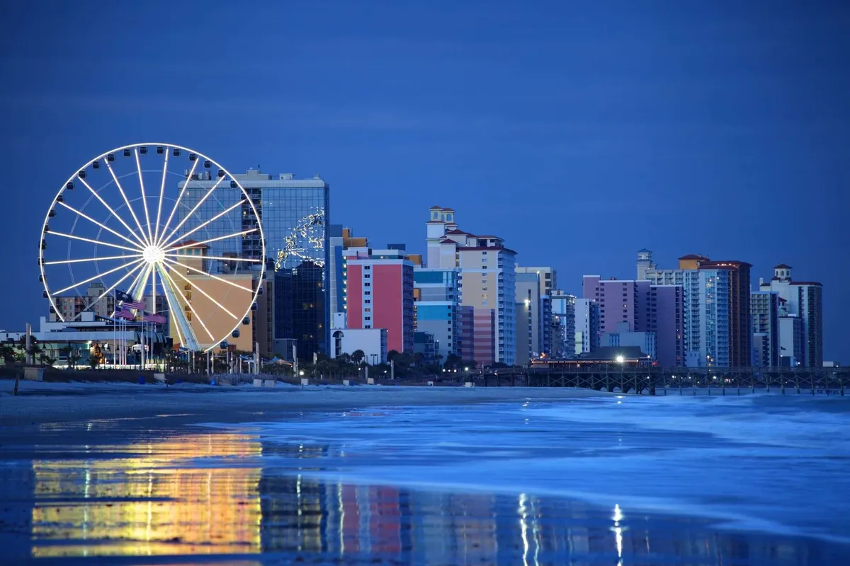 A waterfront scene featuring a ferris wheel and brightly colored buildings, their reflections shimmering on the water. The area is bustling with activity—people stroll along the waterfront, enjoy amusements, and gather at waterside restaurants as the evening lights come on.