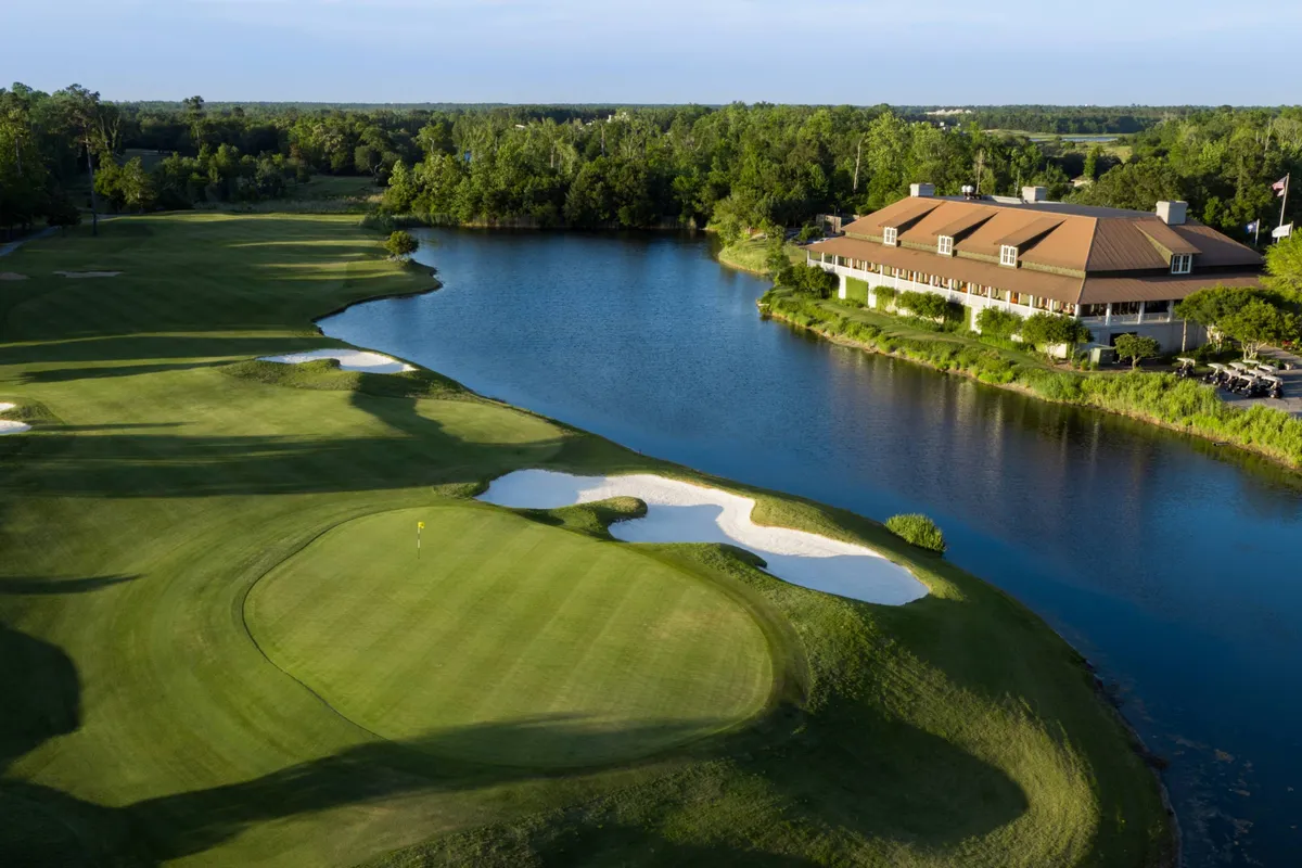 An expansive golf green bordered by a gleaming water body, elegant sand traps, and a stately clubhouse nestled among trees. The tranquil mood and open vista reflect both leisure and the prestige of championship-golf access.