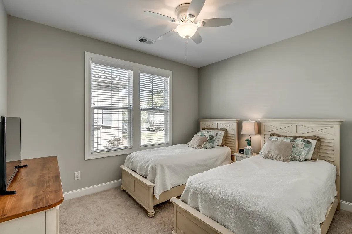 A cozy bedroom with two twin beds, a window with blinds, ceiling fan, and a television on the dresser. The room is bathed in soft natural light, creating a comfortable and tranquil space ideal for relaxing or enjoying quiet family time.
