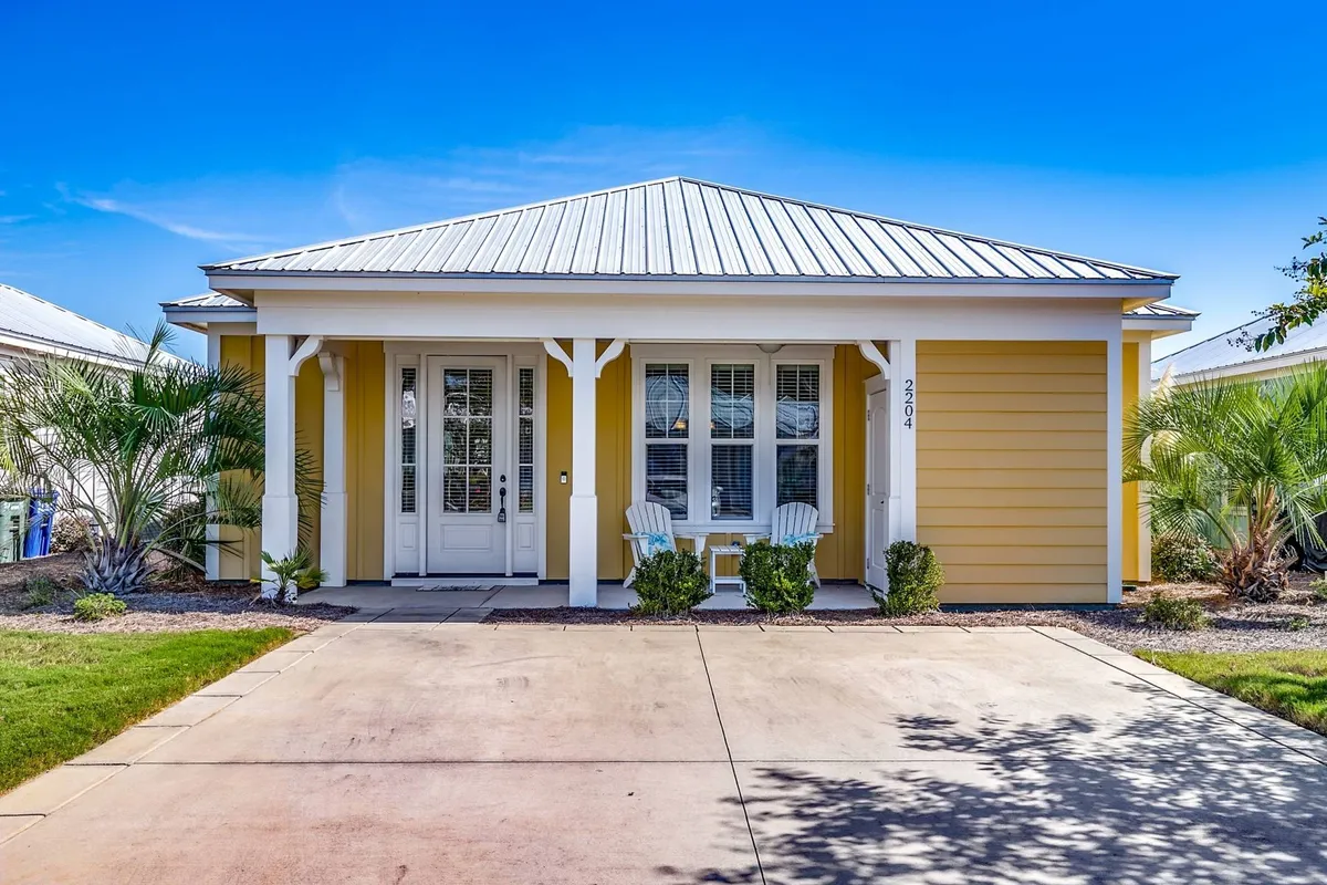 The image features the vibrant yellow exterior of the bungalow with a welcoming front porch, complete with white trim, comfortable rocking chairs, and lush landscaped greenery. The scene radiates a sense of peaceful morning relaxation or a cozy family gathering at sunset.