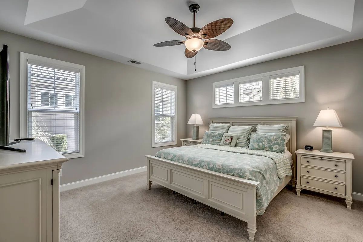This is a serene bedroom with a plush bed adorned with decorative pillows, soft nightstand lighting, a ceiling fan, and large windows with blinds that let in natural morning light. The overall ambiance is calm and restful, with all the elements one might need for a restorative night's sleep.