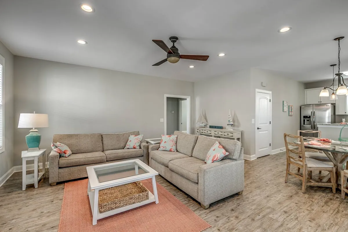A spacious living area featuring comfortable sofas arranged around a coffee table, with a dining table visible in the background. Soft lighting and tasteful decor create a warm, inviting atmosphere, and the layout showcases the open connection between living and dining spaces.