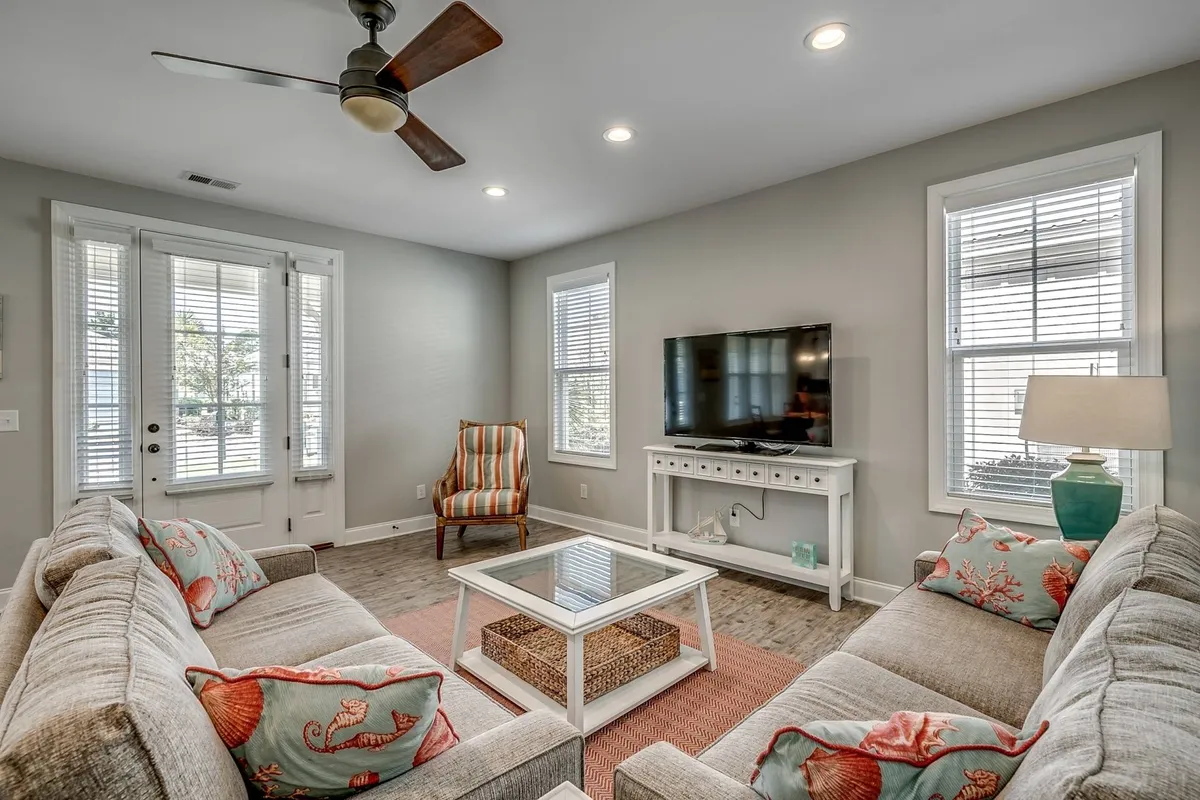 The image showcases a bright, inviting living area with a plush sectional sofa adorned with decorative pillows, a stylish coffee table, and a modern TV console. Sunlight streams in through large windows, creating a warm, welcoming ambiance perfect for relaxation, movie nights, or morning coffee.