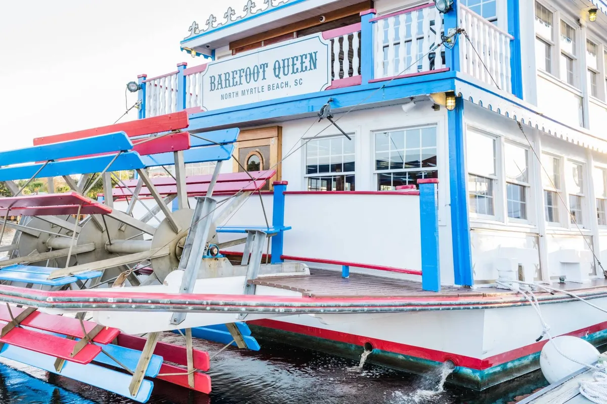 A vibrant, brightly painted paddle wheel boat is moored beside colorful signage, setting a festive mood. Guests can be seen enjoying meals and drinks on deck as the sun sets, with the marina and water providing a lively backdrop.