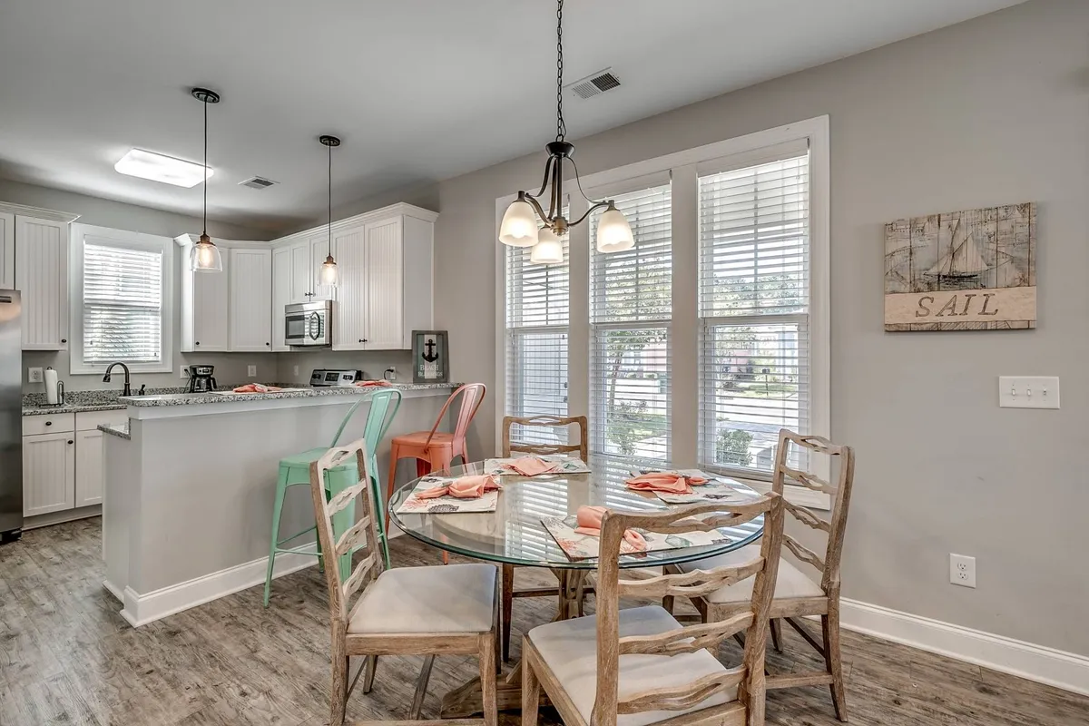A bright, airy dining area with a round glass table surrounded by colorful green and orange chairs. The open kitchen layout can be seen nearby, suggesting a space ideal for family meals, casual gatherings, and shared holiday celebrations.