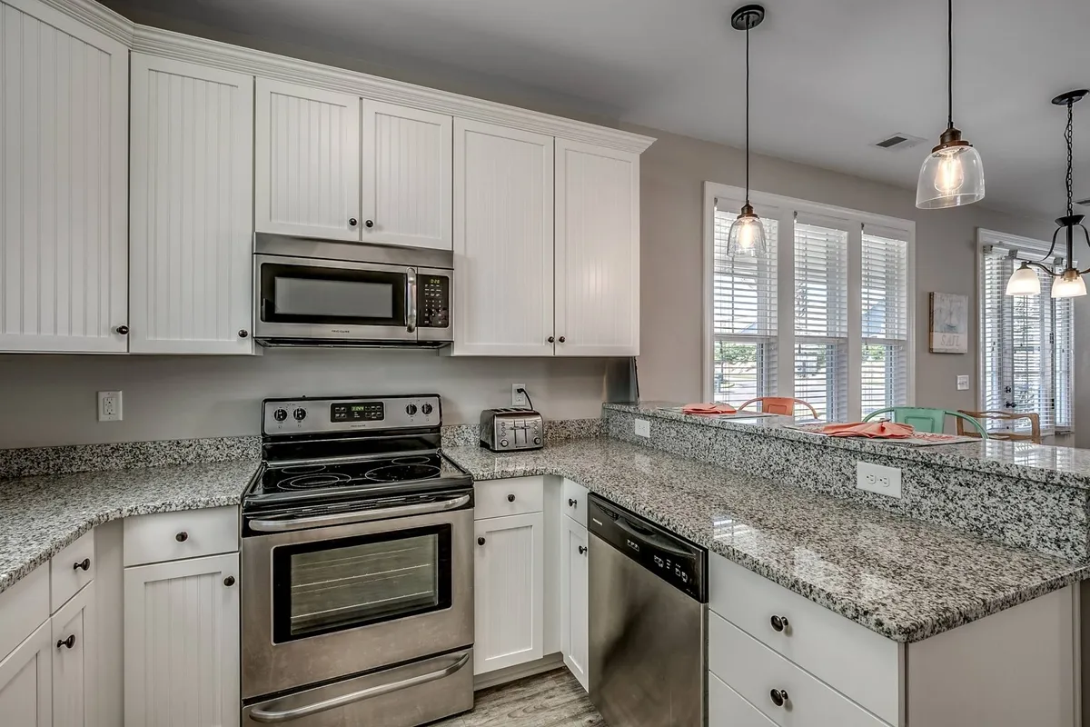 A functional and inviting kitchen space showcasing granite countertops, stainless steel appliances, pendant lighting, and a sunlit window. The area feels welcoming, ready for shared cooking experiences or celebratory meals.