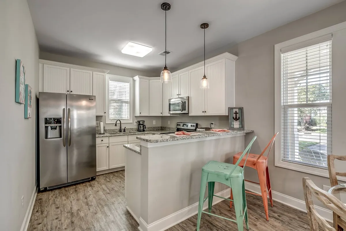 A modern kitchen featuring a stainless steel refrigerator, granite countertops, and a row of colorful stools at the breakfast bar. The space is bright, open, and set up for both meal prep and casual socializing, with appliances and fixtures visible.